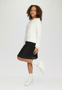 Weißer Sweatshirt mit Grafikdesign, schwarzer plissierter Rock, weiße Sneakers und knielange weiße Socken. Das Model posiert mit einem hochgereckten Bein.