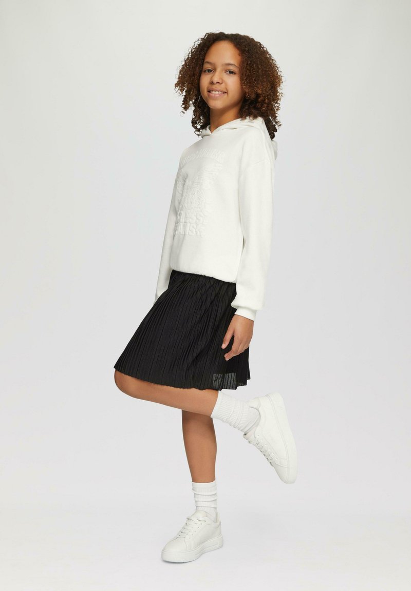 Weißer Sweatshirt mit Grafikdesign, schwarzer plissierter Rock, weiße Sneakers und knielange weiße Socken. Das Model posiert mit einem hochgereckten Bein.