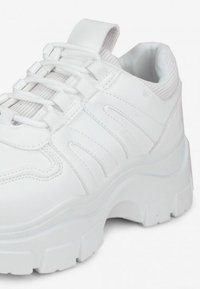 Zapatilla blanca con una parte superior sintética suave, con acentos de malla, cordones redondos y una suela gruesa y texturizada para mayor agarre.