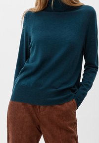 Pull à col roulé teal en tissu doux et texturé, à manches longues et ourlet ajusté. Associé à un pantalon en corduroy côtelé marron.