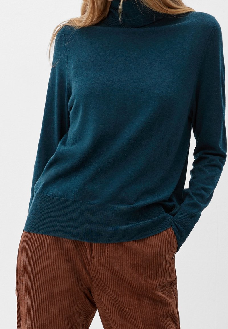 Pull à col roulé teal en tissu doux et texturé, à manches longues et ourlet ajusté. Associé à un pantalon en corduroy côtelé marron.