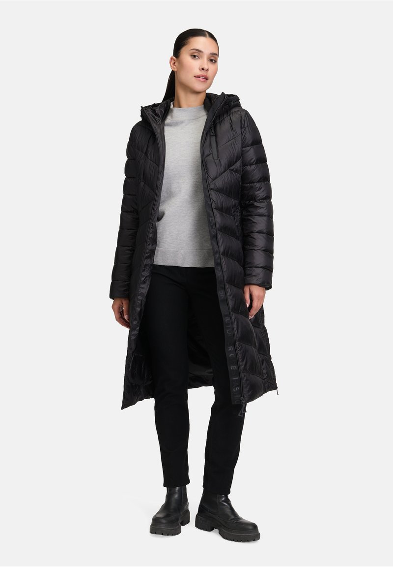 Zwarte pufferjas met een capuchon, met een gestept ontwerp en lange lengte. Gedragen over een grijze trui en gecombineerd met zwarte broek en stevige laarzen.