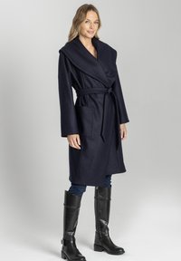 Granatowy wełniany płaszcz z szerokim kołnierzem, paskiem w talii, długimi rękawami i dwiema bocznymi kieszeniami. Modelka w czarnych, sięgających do kolan botkach.