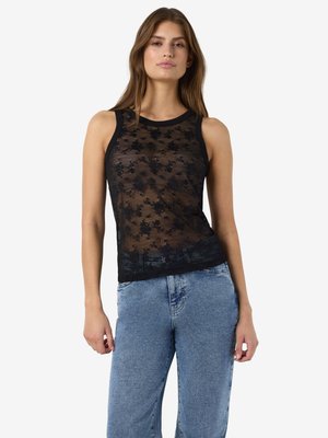 Woman wearing sleeveless black sheer floral lace top and high-waisted blue jeans, standing against plain white background.