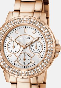 Roségoldenes Metallarmbanduhr mit einem strukturierten weißen Zifferblatt, drei Subzifferblättern und einer Lünette, die mit klaren Kristallen verziert ist. Mit markanten Stundenmarkierungen.