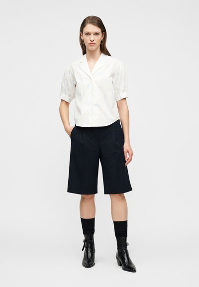 Γυναίκα που φοράει λευκό μπλουζάκι με κοντά μανίκια και διακριτικό φλοράλ σχέδιο, μαύρο σορτς μέχρι το γόνατο, μαύρες κάλτσες και μαύρα μποτάκια, όρθια.