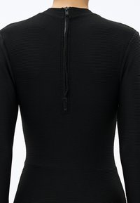 Widok z tyłu osoby noszącej dopasowaną czarną prążkowaną bluzkę z długim rękawem, z widocznym tylnym suwakiem biegnącym od linii szyi do środka pleców.