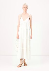 Hvit maxikjole med lagdelte volanger, v-hals og tynne stropper. Modellen holder en liten hvit håndveske og har på seg beige sandaler med stropper.