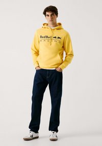 Felpa gialla con cappuccio e logo "Red Bull Racing", abbinata a jeans blu scuro e sneakers bianche. Stile casual, con tasca a marsupio frontale.