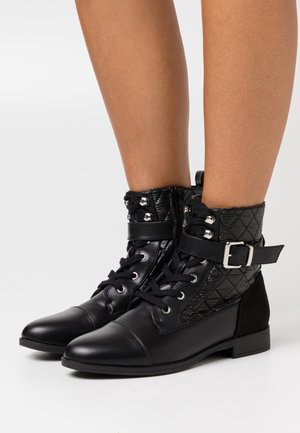 Bottines noires avec un dessus en cuir matelassé, présentant une sangle à boucle, un devant à lacets et un talon plat. Embout lisse et finition mate.