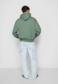Sudadera con capucha verde, combinada con pantalones cargo de un azul claro que cuentan con múltiples bolsillos y un detalle bordado en la parte trasera.