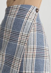 Jupe taille haute à carreaux bleus, blancs et marron, avec un rabat avant fixé par un bouton marron foncé.