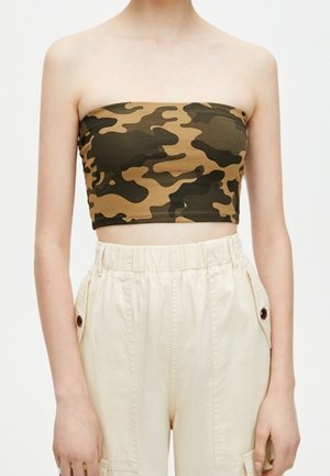 Femme portant un haut court bustier à motif camouflage et un pantalon beige taille haute avec poches latérales et boutons, sur un fond uni.