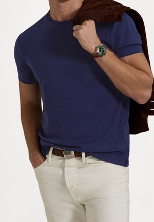 Mann trägt ein marineblaues T-Shirt und weiße Hose, hält eine braune Jacke über der Schulter, mit einer Armbanduhr am linken Handgelenk.