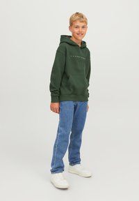 Sudadera verde con bolsillo frontal, corte holgado, puños acanalados. Combinada con jeans azules y zapatillas blancas. Diseño simple y casual.