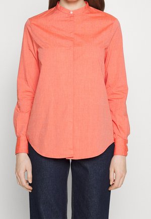 Kvinde iført koralrød langærmet skjorte med knapper og mandarinkrave samt mørkeblå bukser, stående mod en hvid baggrund.