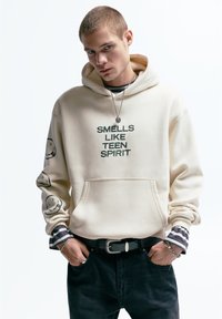 Beige hoodie met de tekst "SMELLS LIKE TEEN SPIRIT" in het groen. Voorzien van smiley-gezicht graphics op de mouwen, de voorzak en een gelaagd gestreept shirt eronder.