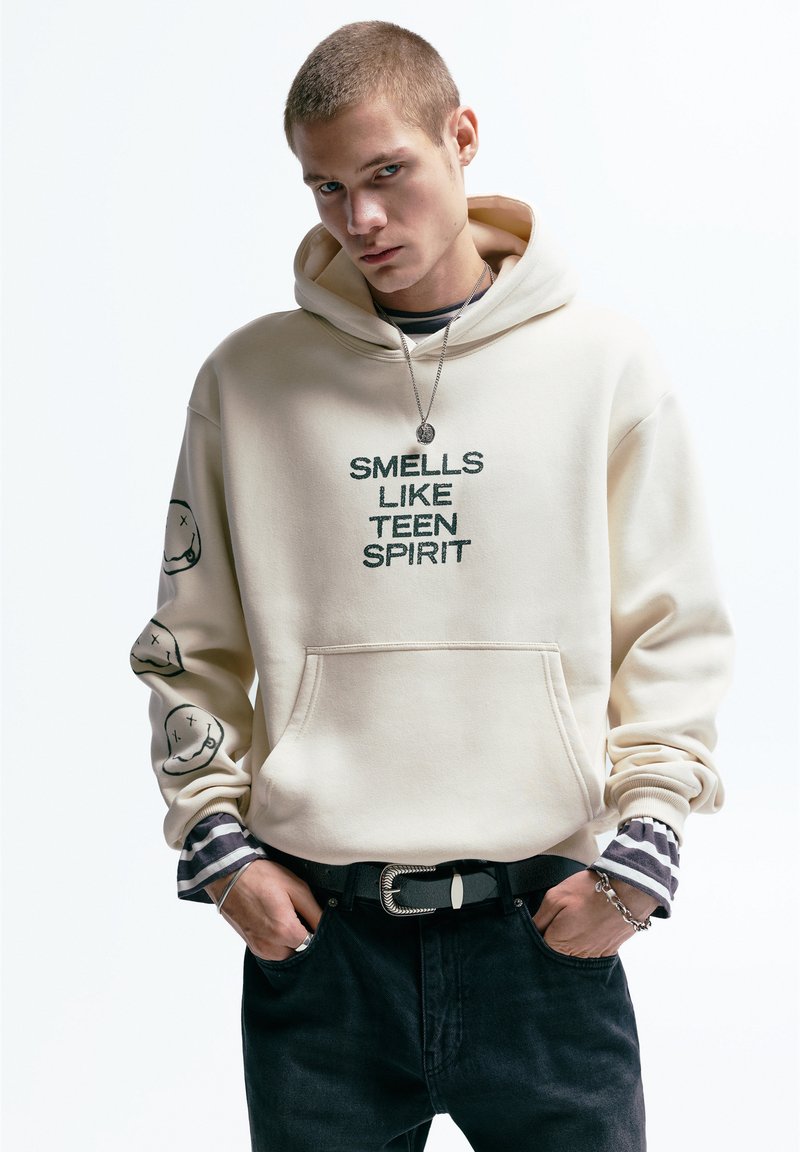 Beige hoodie met de tekst "SMELLS LIKE TEEN SPIRIT" in het groen. Voorzien van smiley-gezicht graphics op de mouwen, de voorzak en een gelaagd gestreept shirt eronder.