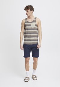 Stribet tanktop i beige og mørkebrun, med en lomme. Sættet inkluderer marineblå shorts, hvide strømper og brune sandaler.