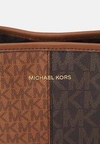 Portefeuille en tissu à motif marron et brun foncé avec cuir, affichant le logo doré "MICHAEL KORS" sur le devant. Détail de surface texturée.