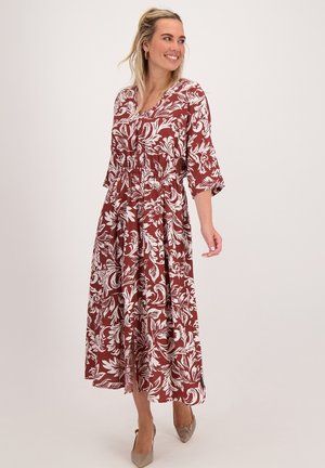 Vrouw in een roestkleurige en witte bloemen midi-jurk met 3/4 mouwen, lachend en naar de zijkant kijkend, met beige puntige hakken.