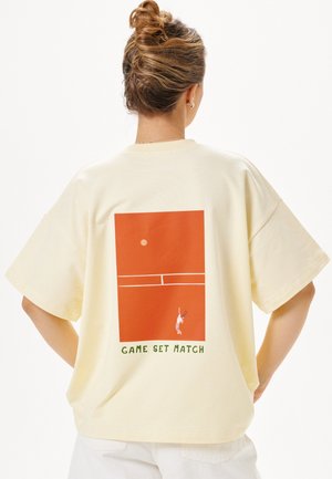 Person mit Dutt, die ein cremefarbenes T-Shirt mit einer Illustration eines roten Tennisplatzes und dem Text "GAME SET MATCH" auf dem Rücken trägt.