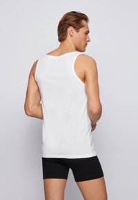 White cotton tank top with wide shoulder straps, fitted design, and a smooth texture, worn with black shorts, viewed from the back.