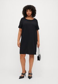 Robe noire à manches courtes avec une coupe décontractée, encolure arrondie, tissu lisse et longueur au genou. Portée avec des chaussures à talons hauts et un petit sac à main.