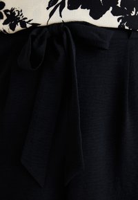 Tejido negro con un acabado texturizado, que presenta un detalle de cintura anudada. La capa superior tiene un patrón floral en blanco y negro.