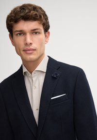 Blazer ribbed blu navy con finitura testurizzata, dotato di una spilla floreale sul revers, un fazzoletto bianco nella tasca e una camicia beige chiaro sotto.