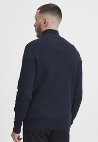 Marinblå stickad tröja med hög krage, ribbade muddar och nederkant, slät textur, har en åtsittande silhuett och minimalistisk detaljering.