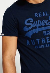 Granatowa bawełniana koszulka z dużym niebieskim napisem "REAL Superdry AUTHENTIC REGISTERED" z przodu oraz metką z logo na rękawie.