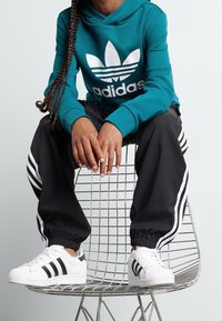 Teal hoodie with white Adidas logo, black pants with white stripes, and white sneakers with black accents, seated on a wire chair.