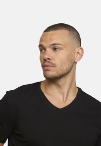 Homme aux cheveux courts et à la barbe de trois jours portant un t-shirt noir à col en V, regardant vers sa gauche contre un fond clair et uni.
