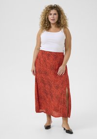 Jupe maxi à motifs rouges avec fentes latérales, fabriquée en tissu léger. Associée à un débardeur blanc simple et des mules noires à talons.