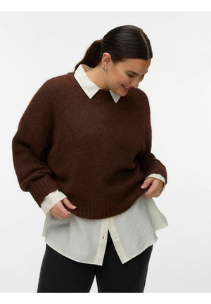 Frau mit dunklem Haar in einem Pferdeschwanz, die einen braunen Pullover über einem weißen Hemd mit Knöpfen und schwarze Hosen trägt, nach unten schaut und lächelt.