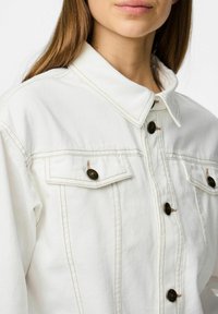 Vit denimjacka med skjortkrage, två bröstfickor med knapplås och kontrasterande bruna sömmar.