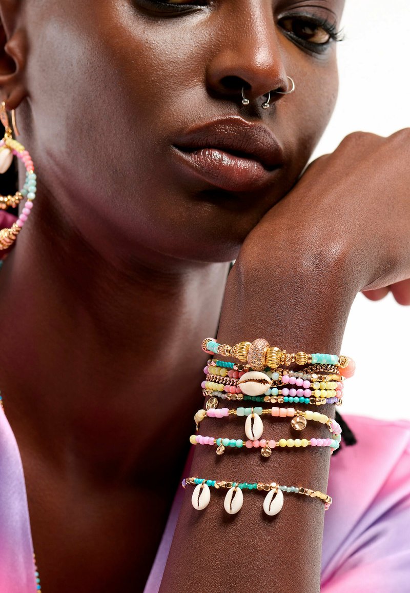 Bracelets colorés en perles avec des accents dorés et des coquillages cowrie sur un poignet à la peau foncée, featuring des perles pastel en rose, bleu et jaune.