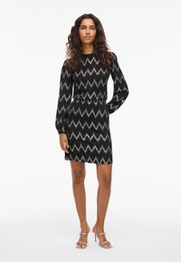 Rochie cu model zigzag negru și argintiu, cu mâneci lungi bufante, talie strâmtă și fustă până la genunchi. Pantofi argintii cu barete completează ținuta.