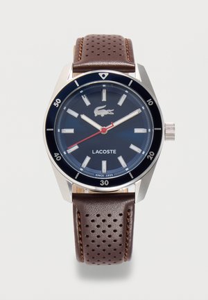 Okrągły srebrny zegarek z ciemnoniebieską tarczą, białymi oznaczeniami godzin, czerwoną wskazówką sekundową, logo Lacoste i perforowanym brązowym skórzanym paskiem.