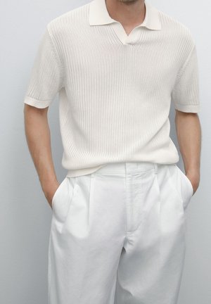 Homme portant un polo en maille blanche à manches courtes, rentré dans un pantalon plissé blanc taille haute, les mains dans les poches.
