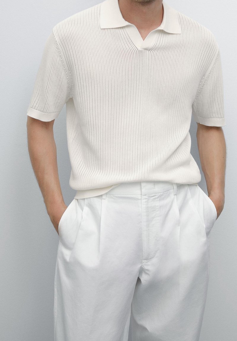 Homme portant un polo en maille blanche à manches courtes, rentré dans un pantalon plissé blanc taille haute, les mains dans les poches.