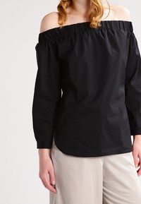 Blusa nera off-the-shoulder con maniche lunghe, scollo elasticizzato e un tessuto liscio e leggero, abbinata a pantaloni beige.