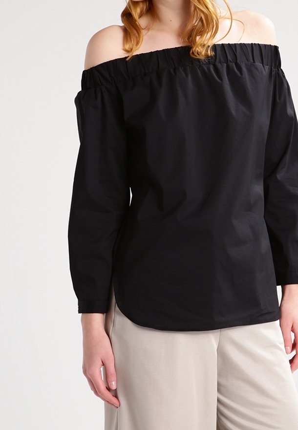 Blusa nera off-the-shoulder con maniche lunghe, scollo elasticizzato e un tessuto liscio e leggero, abbinata a pantaloni beige.