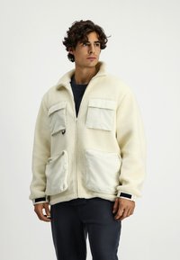 Kremowa polarowa kurtka z wysokim kołnierzem, z dwiema kieszeniami na klatce piersiowej oraz dwiema dużymi kieszeniami z przodu, udekorowana gładkimi, białymi akcentami z tkaniny.