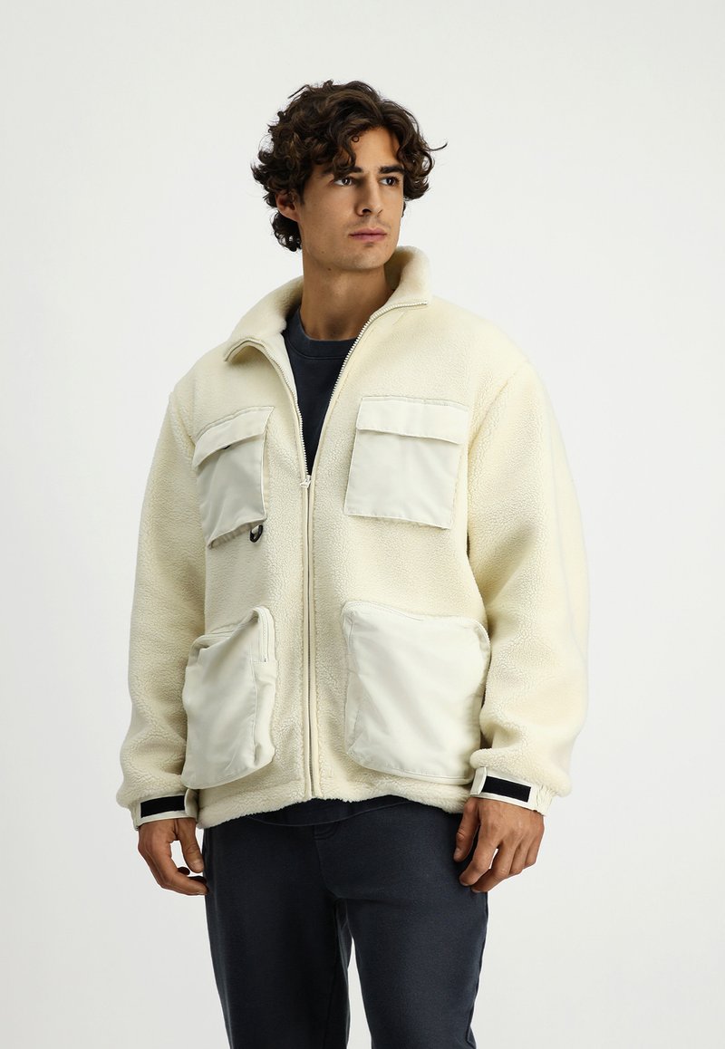 Kremowa polarowa kurtka z wysokim kołnierzem, z dwiema kieszeniami na klatce piersiowej oraz dwiema dużymi kieszeniami z przodu, udekorowana gładkimi, białymi akcentami z tkaniny.
