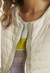 Beżowa pikowana kurtka z metalicznym ozdobnym zamkiem błyskawicznym i teksturowaną złotą lamówką, zestawiona z wielokolorową bluzką w poziome paski.