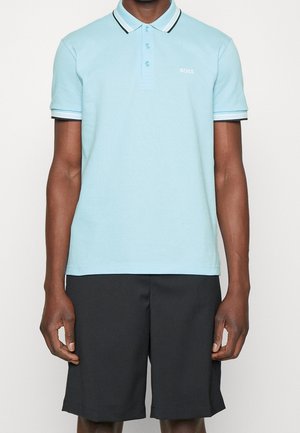 Homme portant un polo bleu clair avec des bordures blanches et un short noir, debout devant un fond uni.