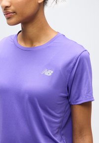 Kvinne iført en kortermet, lilla New Balance treningsskjorte med ribbet tekstur og logo på brystet mot en ensfarget bakgrunn.