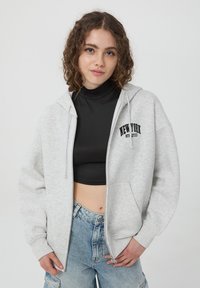 Felpa grigia con zip e tasca frontale, con testo "NEW YORK" in nero. Indossata sopra un top nero corto, accompagnata da un paio di pantaloni corti in denim larghi.